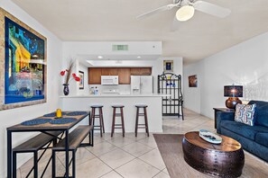 Eine moderne Küche mit weißen Geräten, ein Essbereich mit einem Holztisch und Hockern sowie ein Wohnbereich mit einem blauen Sofa und Zierkissen.