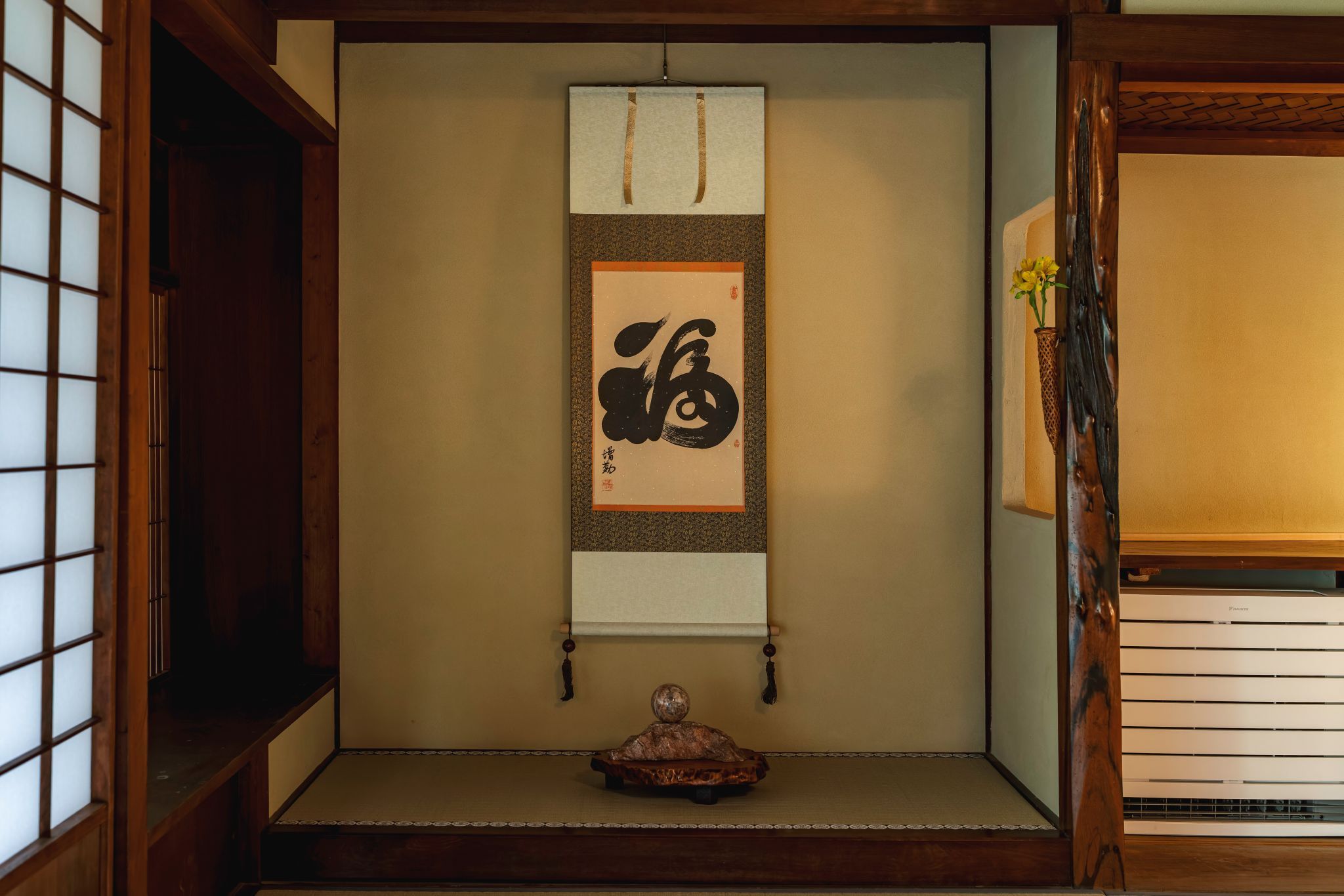 A traditional Japanese room with tatami flooring, a shoji screen, a hanging scroll with calligraphy, and a small wooden sculpture.