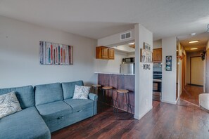 A modern living room with a sofa, a kitchen area with cabinets and appliances, and a hallway leading to another room. - Condo close to hiking trails and base area (Angel Fire)