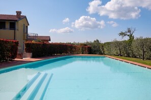 Ein Swimmingpool mit glasklar blauem Wasser, umgeben von einer gepflegten Hecke, ein Haus mit Balkon und ein klarer Himmel.