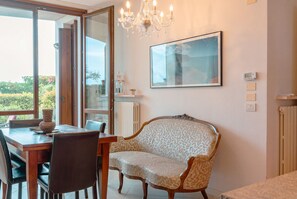Ein Essbereich mit Tisch, Stühlen, einer Couch, einer Kronleuchter, einem gerahmten Bild und Blick durch eine Glastür.