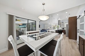 Eine moderne Küche mit einer zentralen Kochinsel, einem Essbereich mit einem Glastisch und Blick durch eine Glastür in einen Wohnraum.
