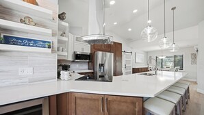 Une cuisine moderne dotée d’un grand îlot central, d’appareils électroménagers en acier inoxydable et de luminaires suspendus.