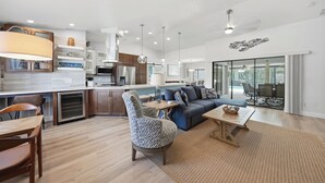 Une cuisine moderne dotée d’un îlot central, un coin repas avec une table et des chaises en bois, un salon avec un canapé et une table basse, et une grande porte-fenêtre coulissante donnant sur un espace extérieur.
