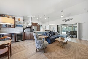 Eine moderne Küche mit Kochinsel, ein Essbereich mit Holztisch und Stühlen, ein Wohnzimmer mit Sofa und Couchtisch sowie eine große Glasschiebetür, die zu einem Außenbereich führt.