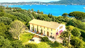 Una villa de estilo mediterráneo con tejado de baldosas, rodeada de exuberante vegetación y con vistas a una bahía costera con barcos.