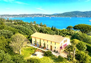 A Mediterranean-style villa with a tiled roof, surrounded by lush greenery and overlooking a coastal bay with boats. - SUPERB MEDITERRANEAN VILLA 12 Pers SEA VIEW & ST TROPEZ, close to BEACHES & GOLF (Grimaud)