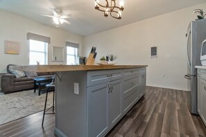 Una cocina moderna con gabinetes grises, encimera de madera y una isla integrada. Incluye un ventilador de techo, un refrigerador y un área de comedor con sofá y mesa.