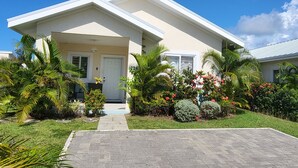 Une maison à deux étages avec un porche à l’avant, un petit jardin avec différentes plantes et fleurs, et un ciel bleu dégagé.