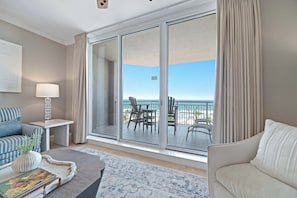 Ein Hotelzimmer mit einer großen Schiebeglasdoor zum Balkon, einem Sofa, einem Couchtisch mit Vase und Büchern sowie einer Lampe.