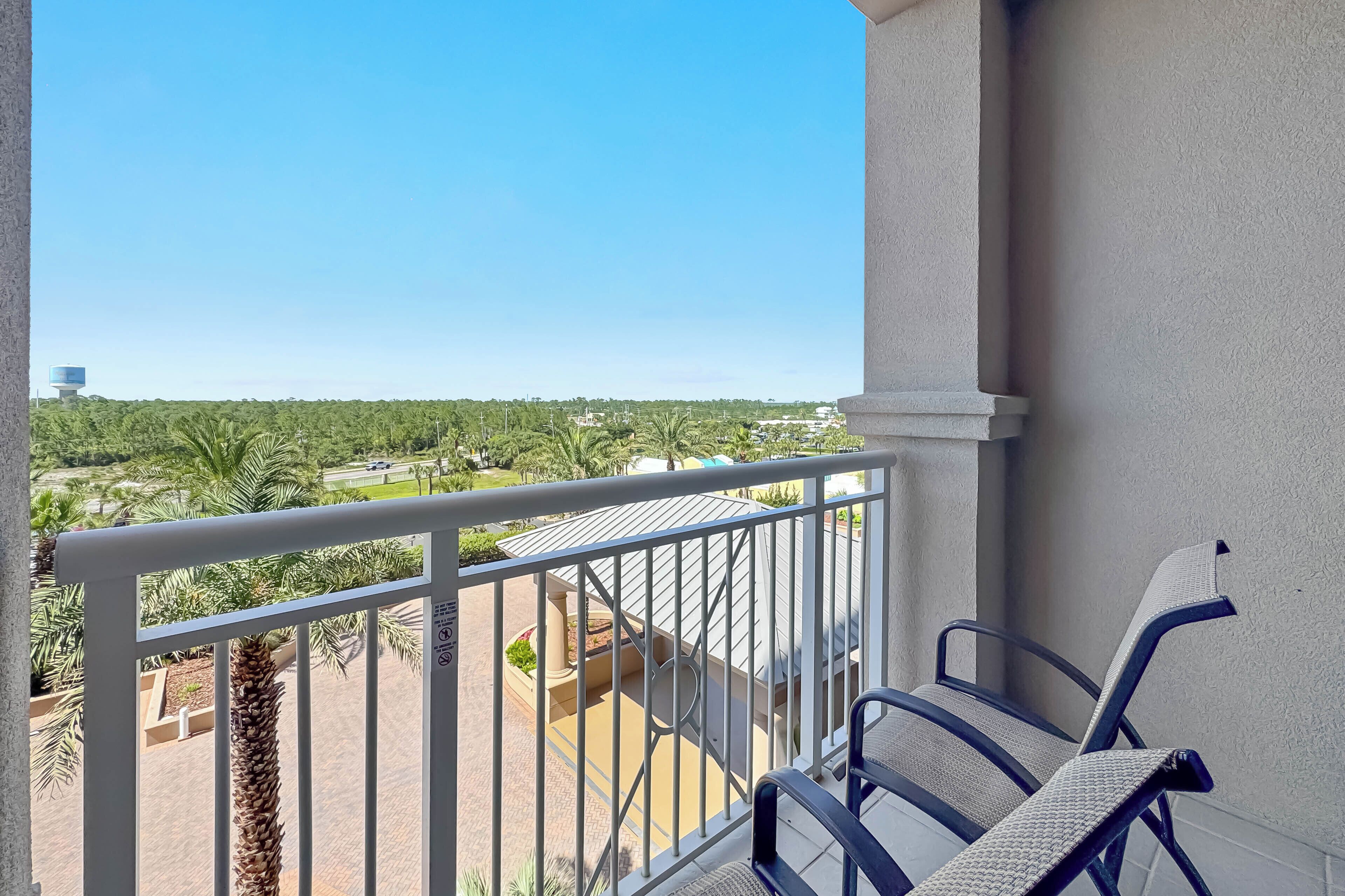 Un balcon doté d’une balustrade blanche, d’une chaise et offrant une vue sur un paysage verdoyant, avec des arbres et un château d’eau.