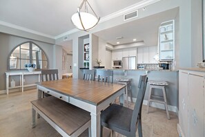 Uma sala de jantar com mesa e cadeiras de madeira, uma cozinha com armários brancos e uma janela grande em arco.