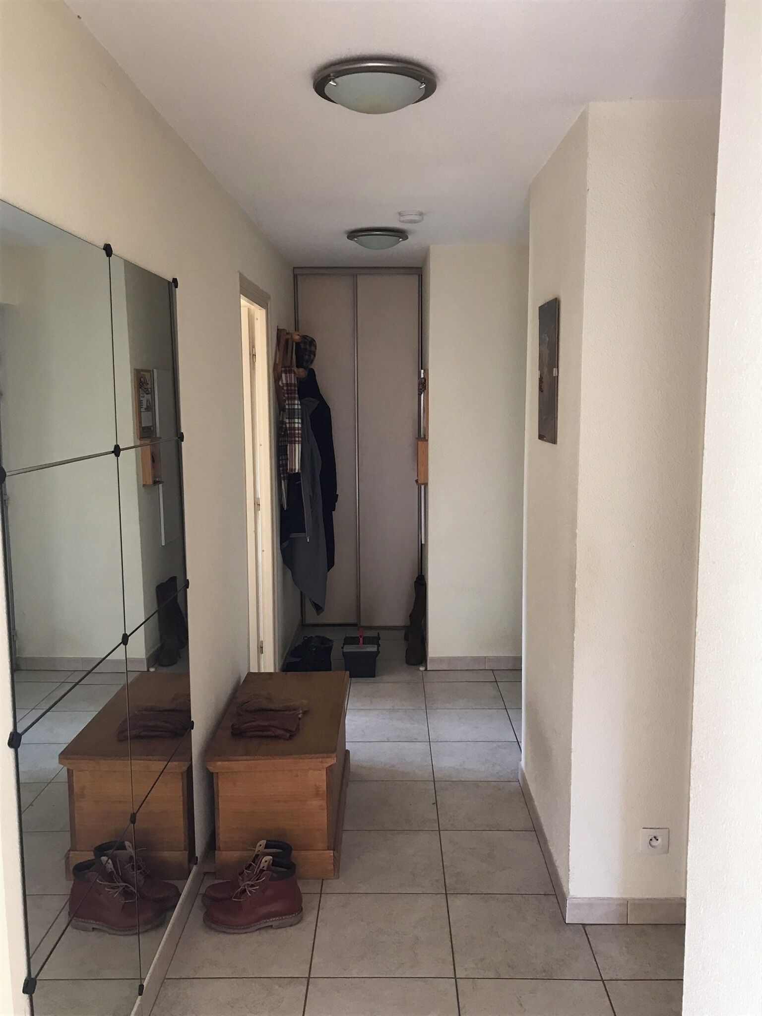 A hallway with a mirror, a coat rack, a wooden bench with shoes, and a door leading to another room.