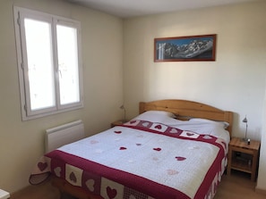 Un dormitorio con cama, ventana, radiador, mesita de noche, lámpara y un cuadro en la pared.