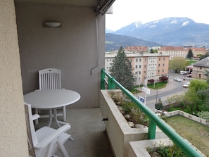 Un balcón con una mesa y sillas, barandal verde y vista a edificios y montañas.