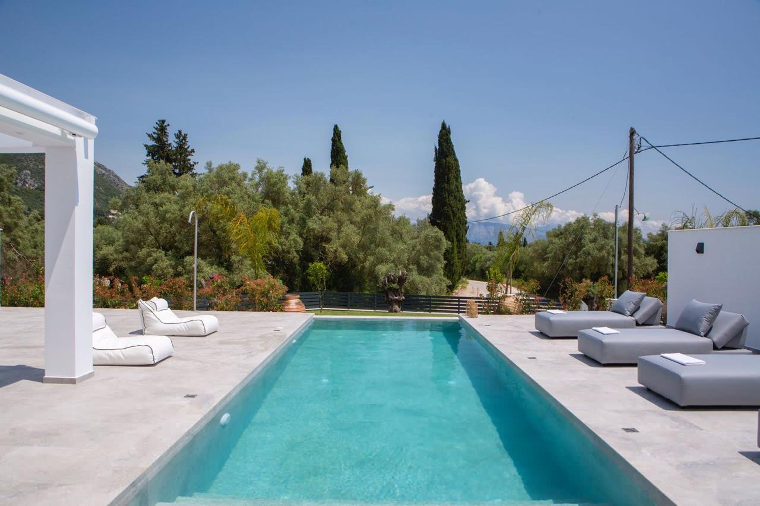 A rectangular pool with white lounge chairs and sofas, surrounded by greenery and trees.