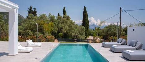 A rectangular pool with white lounge chairs and sofas, surrounded by greenery and trees.