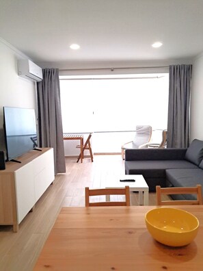 Ein modernes Wohnzimmer mit Sofa, Fernseher, Esstisch und einem großen Fenster.