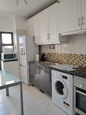 A modern kitchen with white cabinets, a microwave, a refrigerator, a washing machine, and a tiled backsplash. - T2 VISTA MAR in Quarteira (Quarteira)