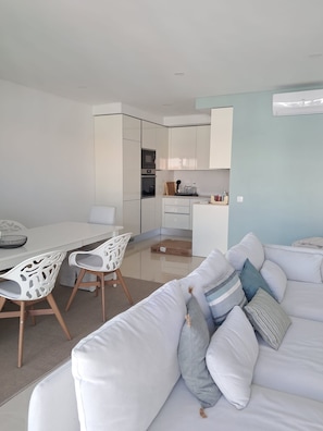 Ein modernes Wohnzimmer mit einem weißen Sofa, einem Esstisch mit Stühlen und einer Küchenzeile mit Schränken und Geräten.