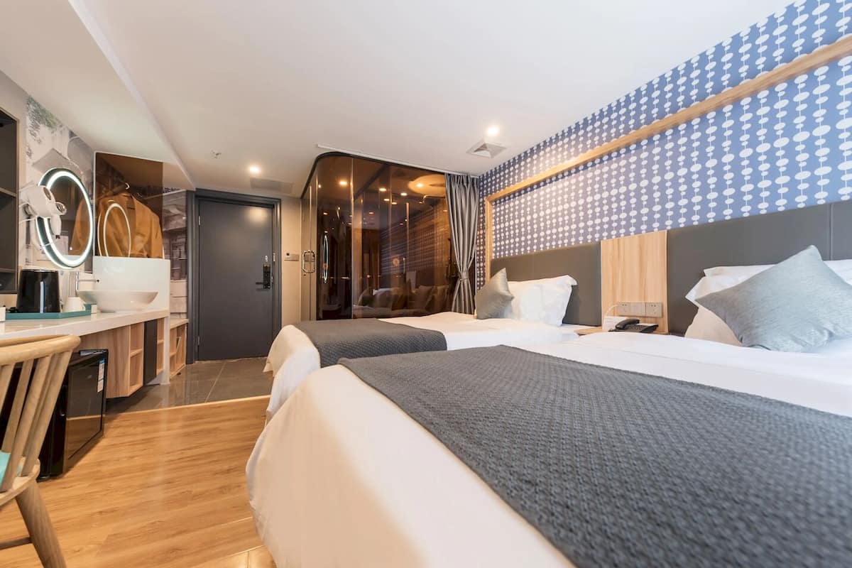A modern hotel room with a desk, two beds, a mirror, and a patterned wall.