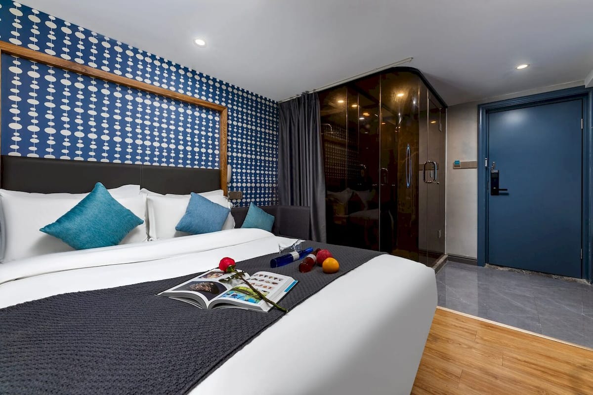 A modern hotel room with a large bed, a dark blue patterned wall, a wooden headboard, and a blue door.