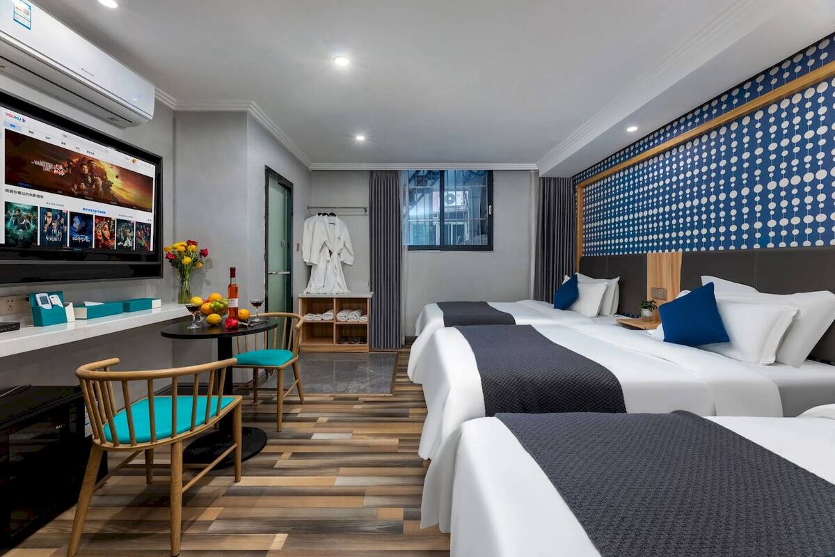 A hotel room with two beds, a desk with a computer, a chair, a small table with a vase of flowers, and a wall-mounted TV.