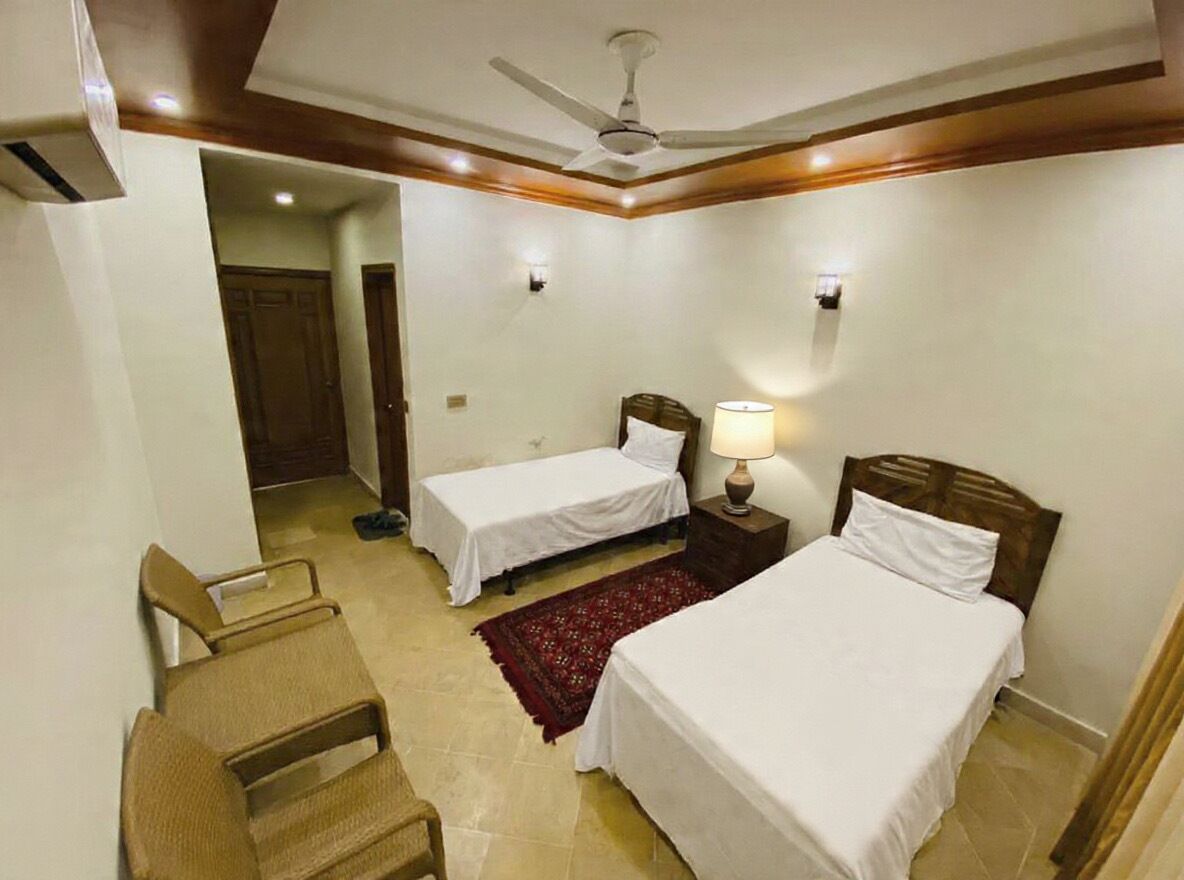 A hotel room with two single beds, a ceiling fan, a nightstand with a lamp, and a red rug.