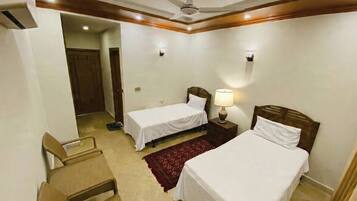 A hotel room with two single beds, a ceiling fan, a nightstand with a lamp, and a red rug.