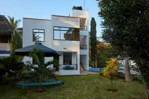 Un bâtiment moderne à deux étages, doté d’un balcon, entouré d’un jardin bien entretenu avec des palmiers et une petite piscine.
