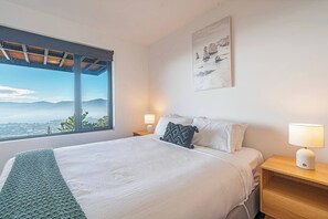 Un dormitorio con una cama grande, dos mesitas de noche con lámparas, vistas amplias a través de un ventanal y un cuadro en la pared.