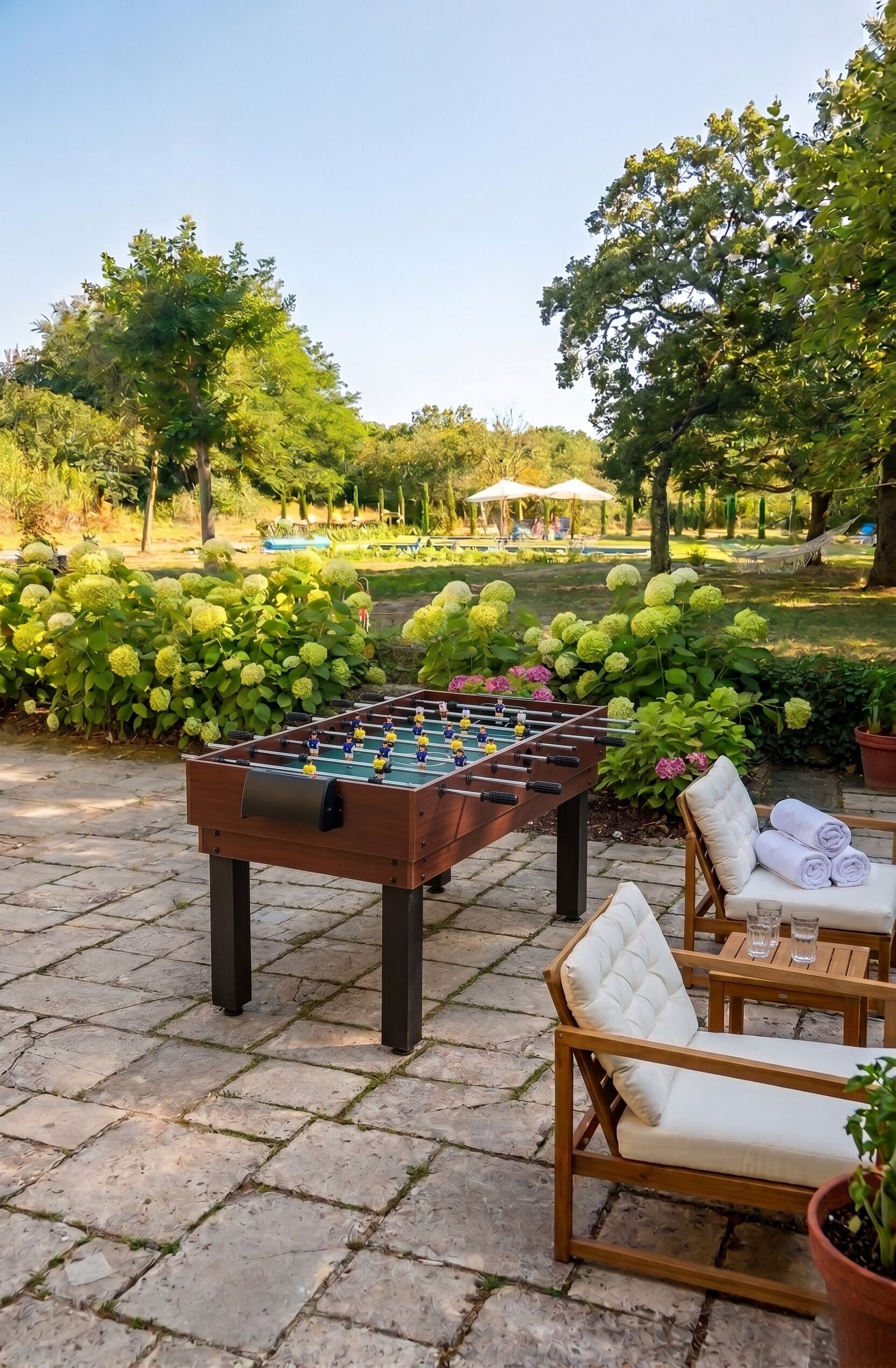Una mesa de fútbol de mesa en un patio exterior con suelo de piedra, rodeada de follaje y una hamaca.