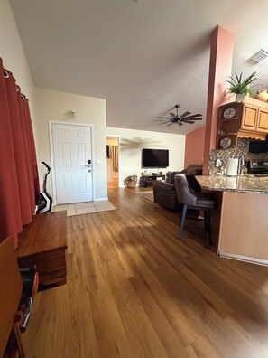 Un'ampia zona giorno con pavimento in legno, un tavolo da pranzo, una cucina con mobili in legno, una ventola a soffitto e una televisione.