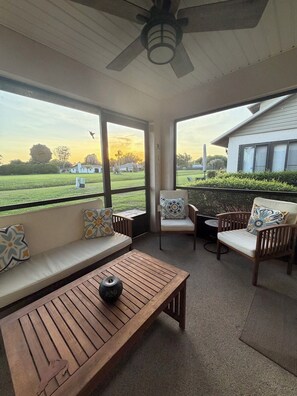A sunroom with a ceiling fan, a wooden table, two cushioned seating areas with patterned pillows, and large windows offering a view of a green landscape and a house. - Beautiful 2 bedroom condo close toGolf courses and the Sebring race track (Sebring)