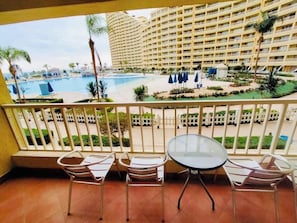 A balcony with a view of a swimming pool, palm trees, and a hotel building.