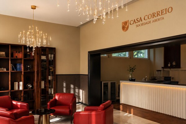A reception area with a chandelier, leather chairs, a bookshelf, and a sign reading 'Casa Correio Sociedade Aberta.'