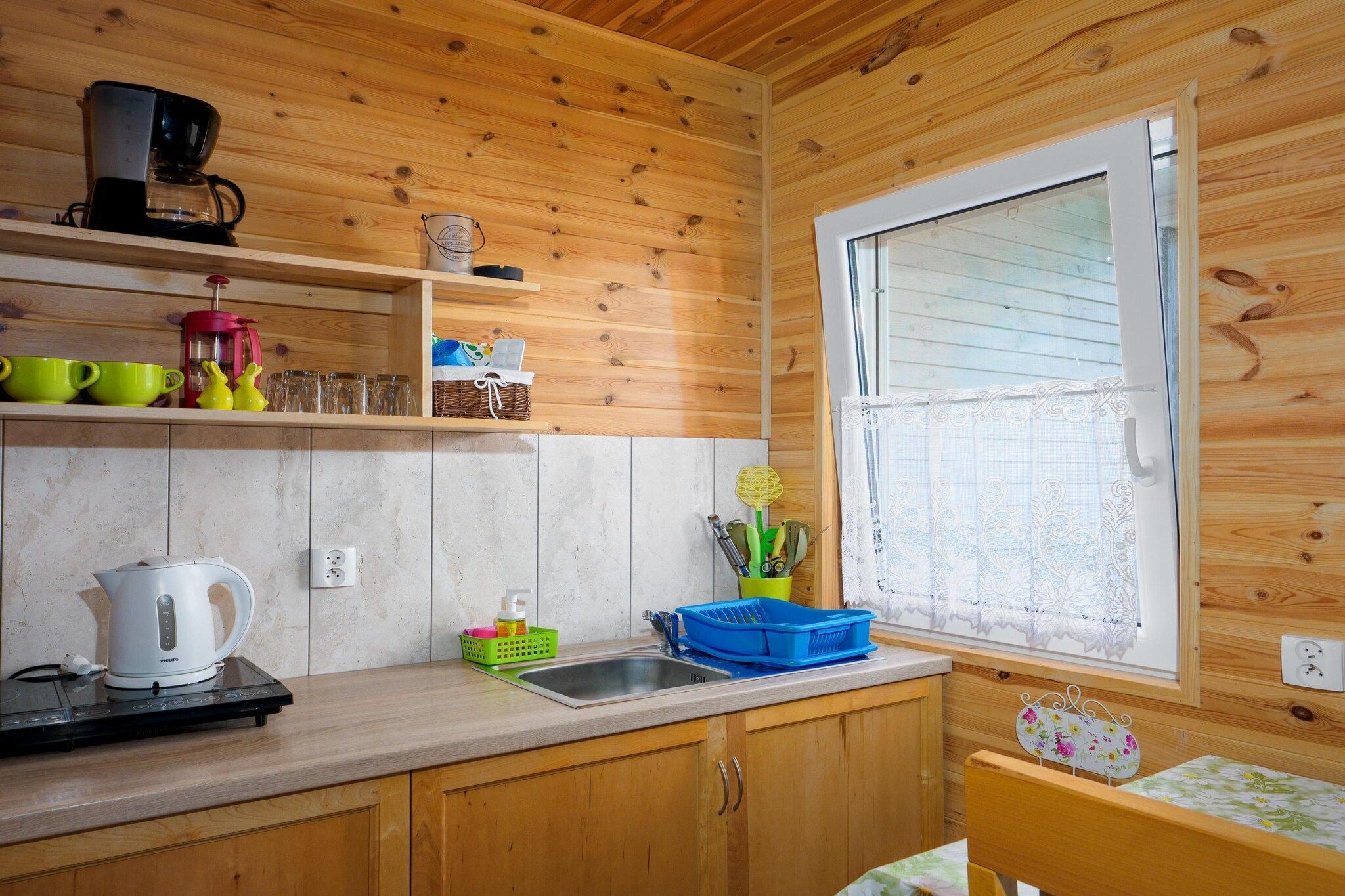 A compact kitchen with wooden walls, a window with lace curtains, a kettle, a coffee maker, and various kitchen utensils.