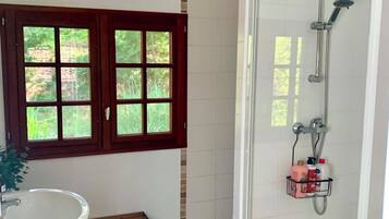 A bathroom with a shower, a sink, a bench, and a window with a view of greenery.