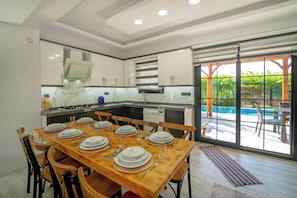 Une cuisine moderne avec des meubles blancs, une table à manger pour six personnes et une vue sur une piscine, visible à travers de grandes portes-fenêtres coulissantes.
