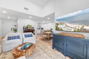 Ein modernes Wohnzimmer mit großem Fernseher, der einen Strandblick zeigt, einem Essbereich mit Holztisch und einer Küche im Hintergrund.