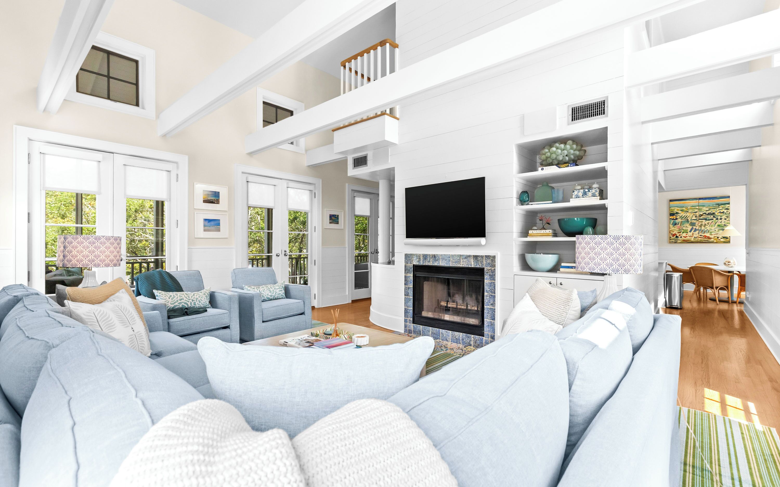 Relax in your spacious living room, where soaring ceilings and soft blue sofas invite you to unwind by the fireplace beneath a wall-mounted TV, with natural light streaming through tall windows.