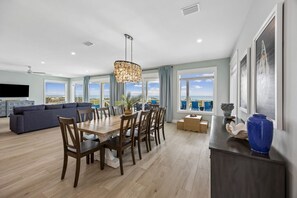 A spacious living area with a dining table, sofas, and a large window offering a view of the sea. - Endless Summer | Brand New Oceanfront Beach House | Hot Tub | Elevator | EV Charger | Sleeps 17 (Fernandina Beach)