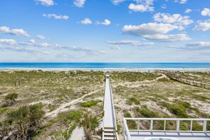 Vista sulla spiaggia con una passerella in legno che si estende verso l'oceano, dune sabbiose e vegetazione rada.
