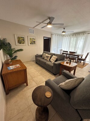 A living room with a sofa, a coffee table, a side table, a ceiling fan, and a dining area with chairs and a table. - Beautiful 2 BR. 2 bath, Sun & Sea Retreat, Rainbow Beach Club Sint Maartan .  (Cupecoy)