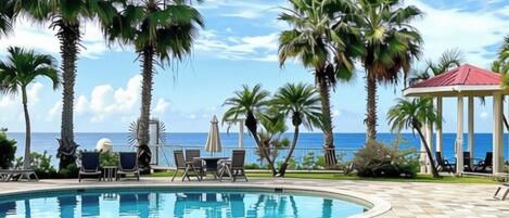 A swimming pool with palm trees and lounge chairs, a gazebo, and a view of the ocean.