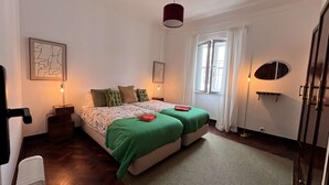 Ein Schlafzimmer mit zwei Betten, einem fensterbelaubten Fenster, einer Hängelampe und Kunstwerken an den Wänden.