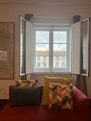 Ein Zimmer mit einem roten Sofa, bunten Kissen, einem großen Fenster mit Vorhängen und Blick auf Gebäude.