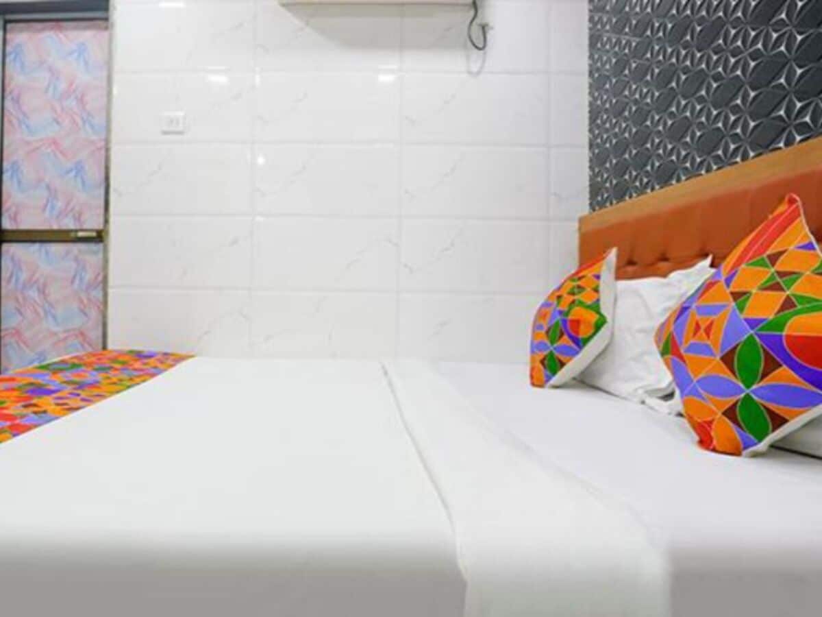 A hotel room with a bed, white bedding, colorful geometric pillows, a white tiled wall, and a textured wall panel.