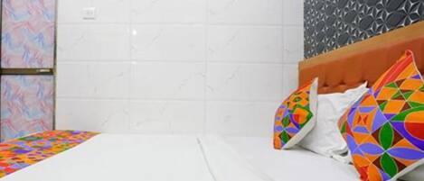 A hotel room with a bed, white bedding, colorful geometric pillows, a white tiled wall, and a textured wall panel.