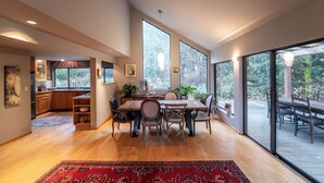 A spacious kitchen with wooden cabinets, a dining area with a large table and chairs, and large windows offering views of the outdoors. - Beautiful Farmhouse & thermal Spa in Forest (Sechelt)